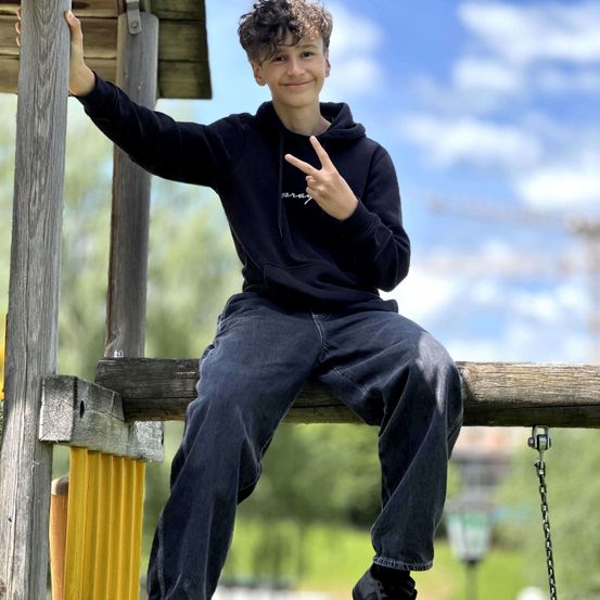 Ein junger Junge in einem schwarzen Hoodie und Jeans sitzt auf einer Holzbalken in einem Park. Er macht ein Friedenszeichen mit einer Hand.