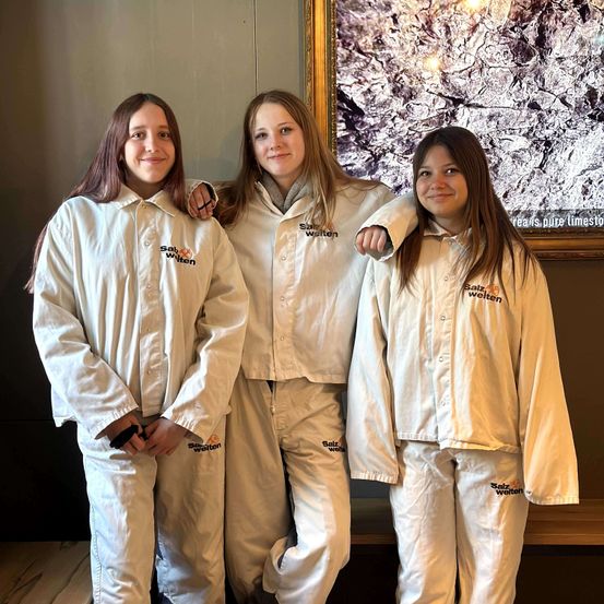 Drei Frauen in passenden weißen Overalls mit 'Salzwelten' bedruckt posieren zusammen in einem Raum mit einer großen Wandkunst dahinter.