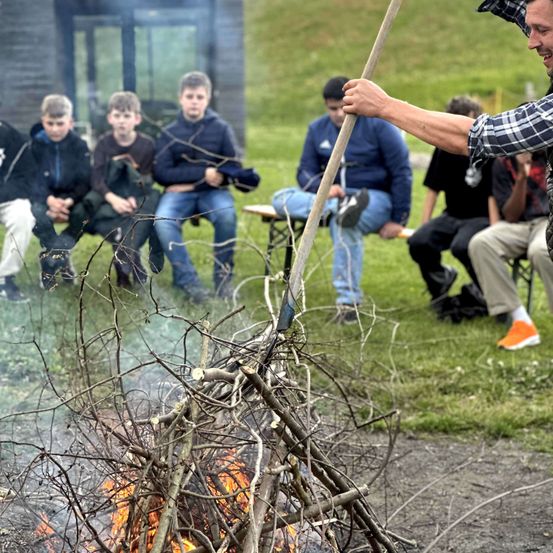 Eine Gruppe von Jungen sitzt um ein Lagerfeuer herum und beobachtet einen Mann mit einem Stock. Sie tragen alle Jacken und Jeans. Der Mann lächelt und scheint mit den Jungen zu sprechen.
