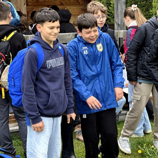 Eine Gruppe junger Jungen und Mädchen steht draußen auf Gras, einer der Jungen trägt einen blauen Mantel mit den Buchstaben AB.