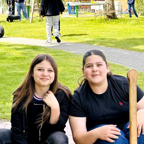 Zwei Mädchen sitzen auf einem Weg in einem Park und lächeln für ein Foto. Hinter ihnen befindet sich ein Rasenbereich mit einem Holzstab und einer Reifenschaukel. Im Hintergrund gehen Leute vorbei.
