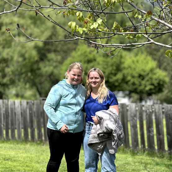 Zwei Frauen, beide lächelnd, stehen in einem Garten. Eine hält eine Jacke, die andere trägt eine hellblaue Kapuzenjacke. Hinter ihnen befindet sich ein Holzzaun und üppige Grünflächen.