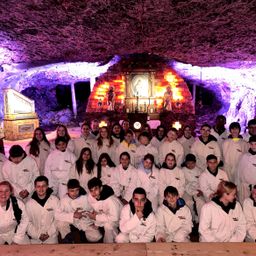 Eine Gruppe junger Menschen in weißen Kitteln posiert für ein Foto in einer Höhle mit einem Altar und religiösen Statuen, die von lila und weißen Lichtern beleuchtet werden.
