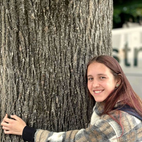 Ein Mädchen mit braunen Haaren umarmt einen Baum und lächelt. Sie trägt eine karierte Jacke. Im Hintergrund ist ein Schild mit weißen Buchstaben zu sehen.