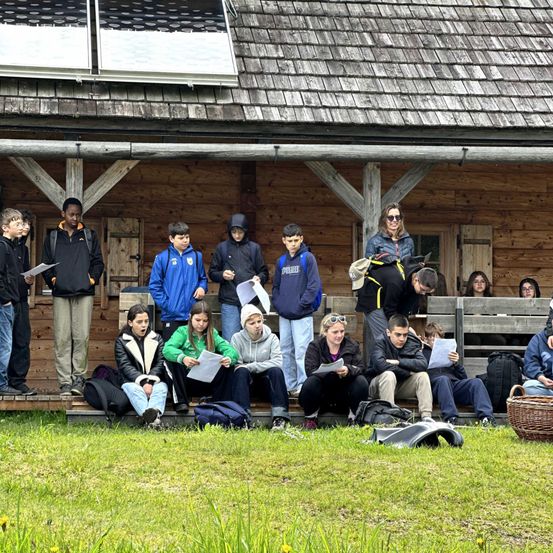 Eine Gruppe von Schülern sitzt auf einer Holzveranda und hält Papiere in den Händen, wahrscheinlich um Notizen zu machen. Einige stehen hinter ihnen. In der Nähe liegen Rucksäcke und ein Korb auf dem Gras.