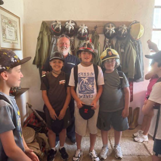 Eine Gruppe von Kindern, einige mit Mützen, stehen vor einer Wand mit Helmen, Jacken und Stiefeln. Ein Mann in einem blauen Hemd lächelt sie an.
