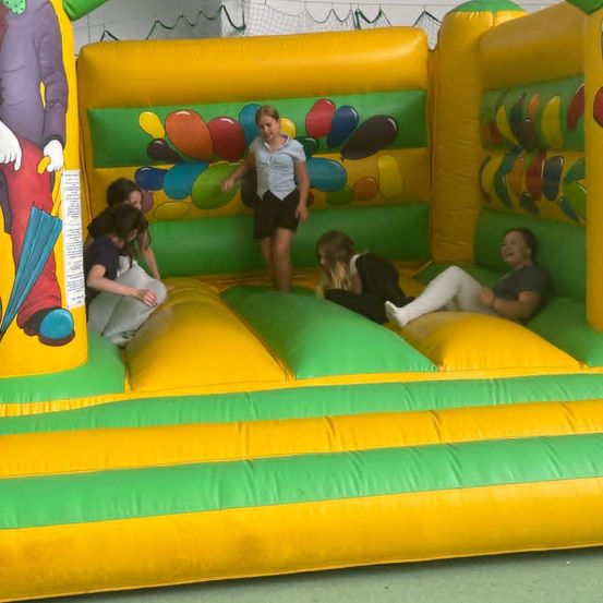 Mehrere Kinder spielen in einem bunten aufblasbaren Hüpfburg mit Luftballons und einem Clown an der Seite. Ein Mädchen steht am Eingang.