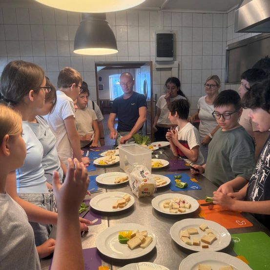 Eine Gruppe von Menschen versammelt sich um einen Tisch mit Sandwiches, Gemüse und Brot. Ein Mann steht vorne und bereitet Essen zu. Sie scheinen eine Kochstunde zu lernen.