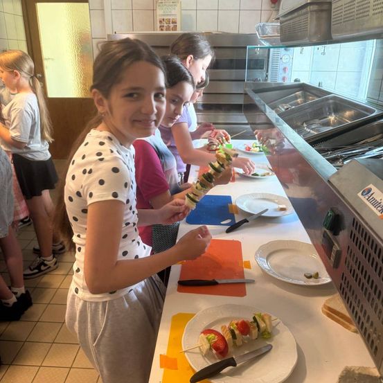 Eine Gruppe von Mädchen ist in einer Küche und bereitet Essen zu. Ein Mädchen macht einen Gemüsespieß. Andere Mädchen bereiten auch Essen zu. Es gibt Teller, Messer und Brot auf dem Tisch.