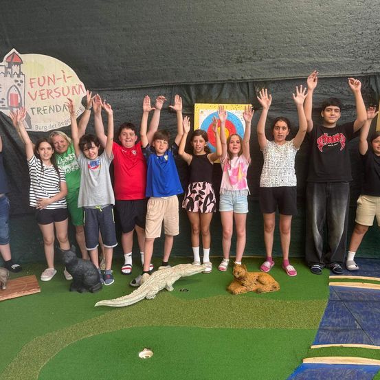 Eine Gruppe von Kindern posiert für ein Foto mit erhobenen Händen in einem Innenbereich mit einem Ziel für Bogenschießen und einer Wand mit dem Text 'Fun-i-Versum Trend'.