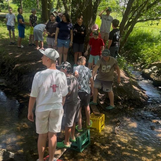 Kinder versammeln sich um einen kleinen Bach, einige treten auf Holzbretter und Kisten. Einige tragen Hüte und Sandalen, andere sind barfuß. Sie beobachten das Wasser.