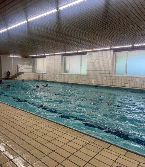 Ein Hallenbad mit Schwimmern. Der Pool hat einen gefliesten Boden und weiße geflieste Wände. Es gibt Glasfenster und eine Holzdecke mit Leuchtstofflampen.