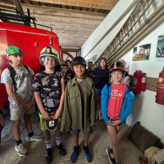 Vier Kinder stehen in einer Feuerwache und tragen Helme. Hinter ihnen steht ein Feuerwehrauto, Leitern und ein Feuerlöscher. Eine Frau steht im Hintergrund.