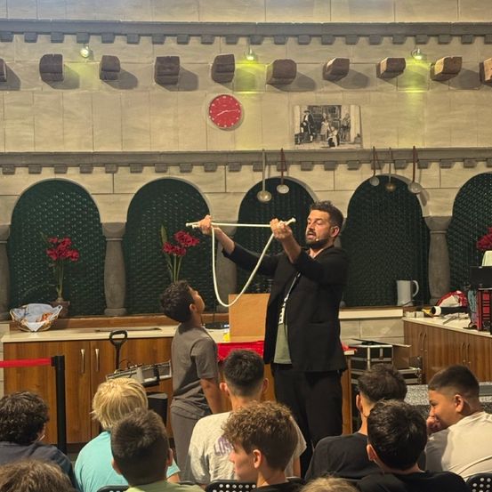 Ein Mann führt einen Zaubertrick mit einem Seil vor einem jungen Publikum vor. Die Szene spielt sich in einem Raum mit Bühne und einer Uhr darüber ab.