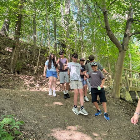 Eine Gruppe junger Leute mit Sonnenbrillen wandert in einem Wald mit einer Holzbrücke im Hintergrund.
