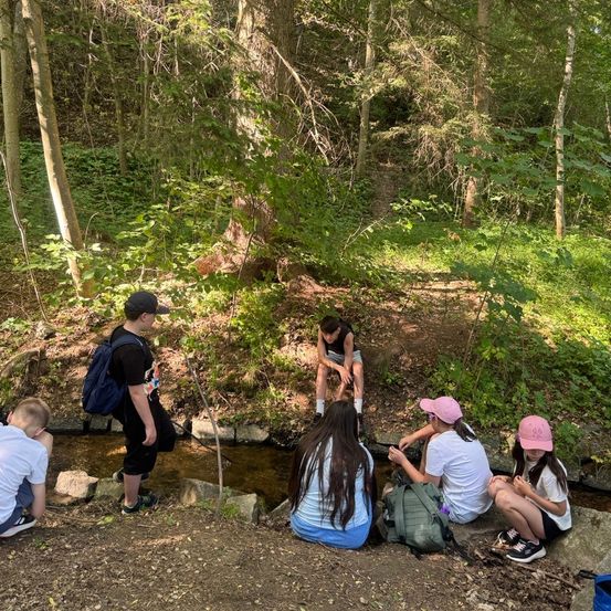 Eine Gruppe von Kindern erkundet ein Waldgebiet. Einer steht und hält einen Stock, während andere an einem Bach sitzen. Einige tragen Hüte, und Rucksäcke sind verstreut.
