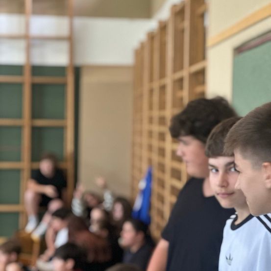 Eine Gruppe von Teenagern sitzt in einem Klassenzimmer. Einige stehen, andere sitzen. Links ist eine Leiter, rechts eine Tafel.