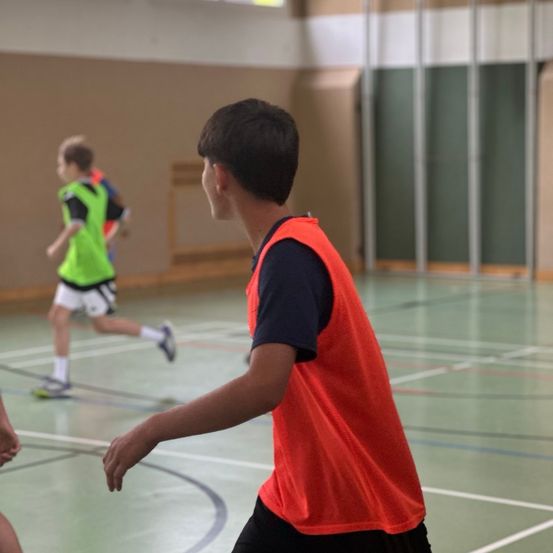 Zwei Jungen spielen Basketball in der Halle. Einer in Grün, der andere in Rot, beide tragen Trikots. Eine Holzbank im Hintergrund.