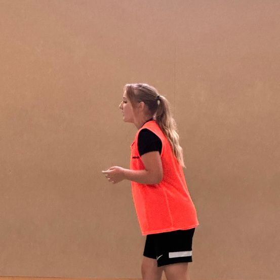 Eine Frau in einem neonorangefarbenen Sicherheitsweste und schwarzen Shorts steht in einem Fitnessstudio.
