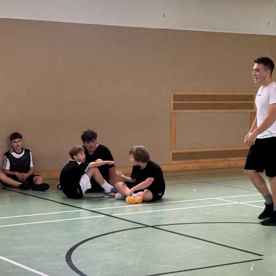 Fünf junge Männer sitzen auf dem Boden eines Hallenspielfeldes und unterhalten sich. Ein Mann steht auf und spricht die Gruppe an. Das Spielfeld hat einen weißen Boden und braune Wände.