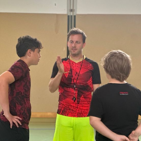 Drei Personen stehen in einer Turnhalle. Der zentrale Mann trägt ein rot-schwarzes Shirt mit Lanyard. Der Mann auf der linken Seite trägt ein rot-schwarzes Shirt. Der Mann auf der rechten Seite trägt ein schwarzes Shirt.