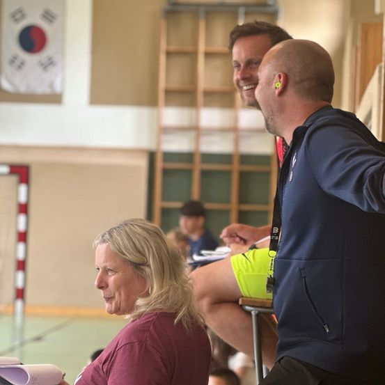 Drei Personen sitzen in einem Fitnessstudio. Eine Frau lächelt und schaut zur Kamera. Zwei Männer lehnen sich zu ihr hinüber. Ein Banner und eine Leiter sind im Hintergrund zu sehen.