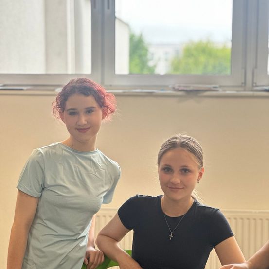 Zwei Frauen posieren für ein Foto in einem Raum mit einem großen Fenster, draußen sind Bäume zu sehen. Die Frau auf der linken Seite hat rote Haare und die Frau auf der rechten trägt eine Kreuzkette.