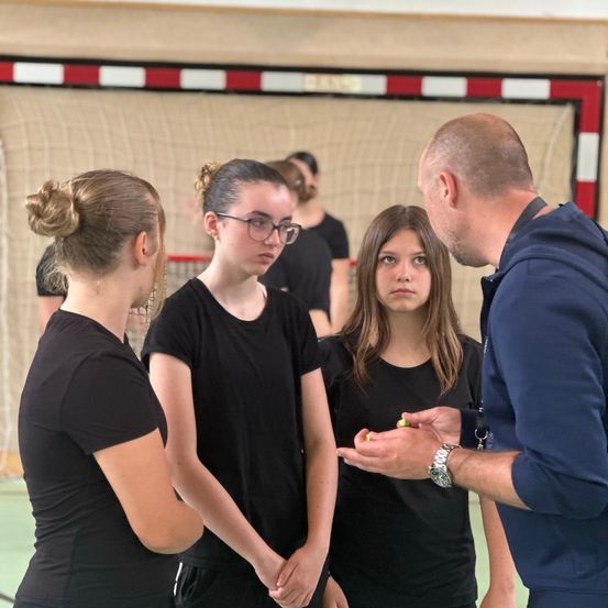 Drei Mädchen und ein Mann stehen in einem Fitnessstudio und schauen sich gegenseitig an. Die Mädchen tragen schwarze T-Shirts und der Mann eine blaue Kapuzenjacke. Er hält ein gelbes Objekt. Im Hintergrund befindet sich ein rot-weißes Netz.