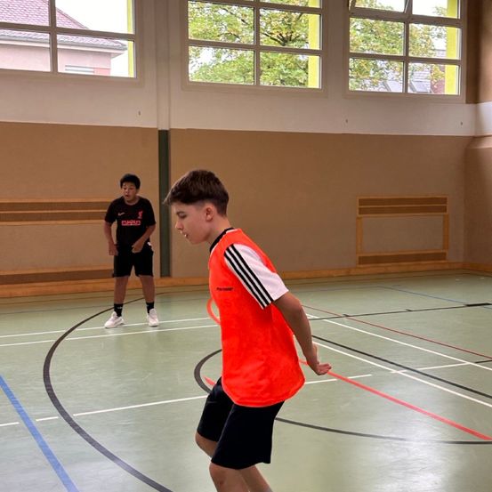 Zwei Jungen spielen Badminton in einer Turnhalle. Einer in einem schwarzen Trikot, der andere in einer orangefarbenen Weste. Sie konzentrieren sich auf das Spiel.