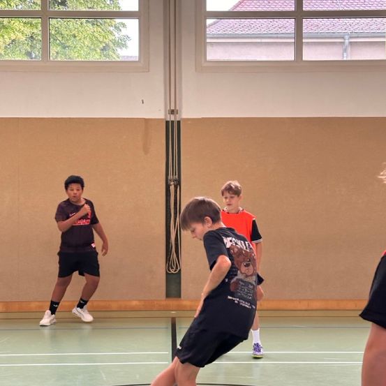 Drei Jungen spielen drinnen ein Spiel. Ein Junge in Schwarz beugt sich nach unten, und zwei andere stehen hinter ihm und beobachten. Der Innenraum hat braune Wände und Fenster.