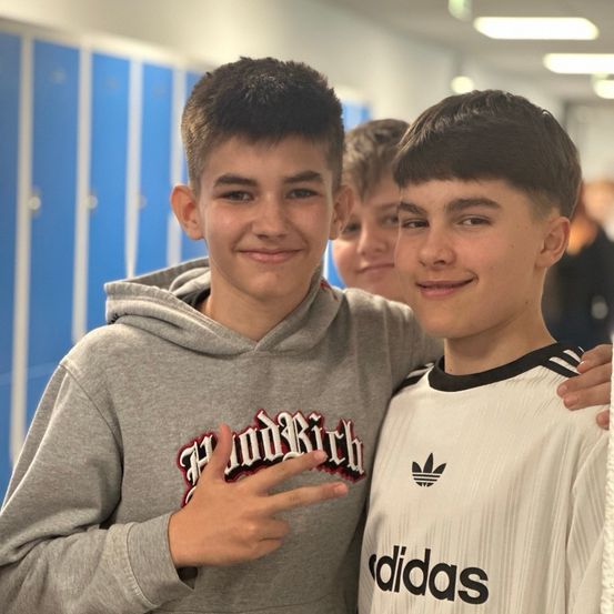 Zwei junge Jungen in Schuluniformen, einer in einem Hoodie und der andere in einem Adidas-Shirt, stehen eng beieinander und lächeln.