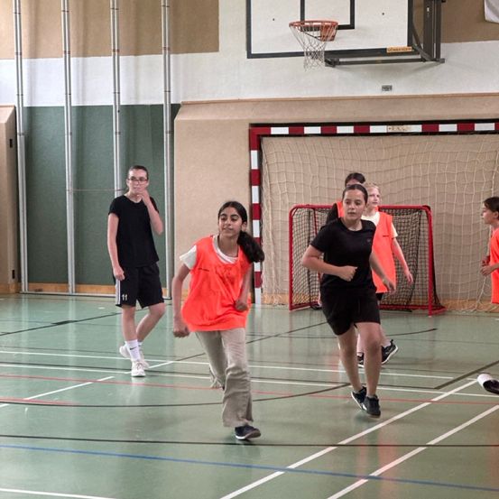 Mehrere junge Menschen laufen auf einem Sporthallenboden mit einem Basketballkorb im Hintergrund. Zwei Mädchen tragen orange Weste.