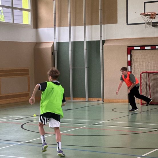 Zwei Jungen spielen eine Runde Dodgeball auf einem Indoor-Spielfeld. Ein Junge läuft auf den Ball zu, während der andere verteidigt.