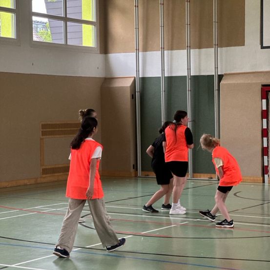 Fünf Personen in orangefarbenen Westen befinden sich in einer Turnhalle, gehen und schauen auf ein Kind, das sich bücken muss. Die Turnhalle hat Fenster und Wände, die in Grün und Beige gestrichen sind.