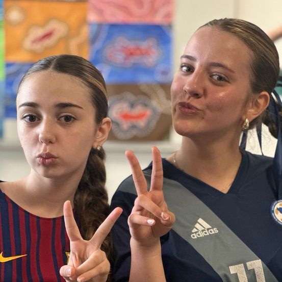 Zwei junge Frauen, eine mit langen Haaren und eine andere mit geflochtenen Haaren, machen Friedenszeichen mit ihren Fingern. Sie tragen Sporttrikots. Ein bunter Hintergrund ist dahinter.