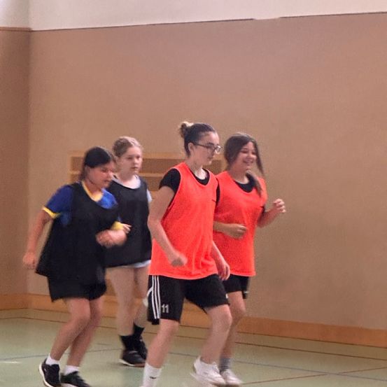 Vier Mädchen in Sportbekleidung, zwei mit orangefarbenen Westen, laufen in einer Sporthalle, möglicherweise üben sie eine Mannschaftssportart.