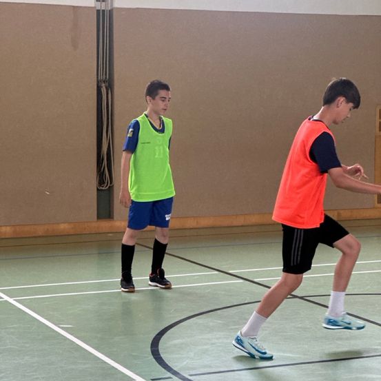 Zwei Jungen spielen Futsal. Der Junge in Grün schaut den Jungen in Orange an. Der Boden hat weiße und schwarze Linien.