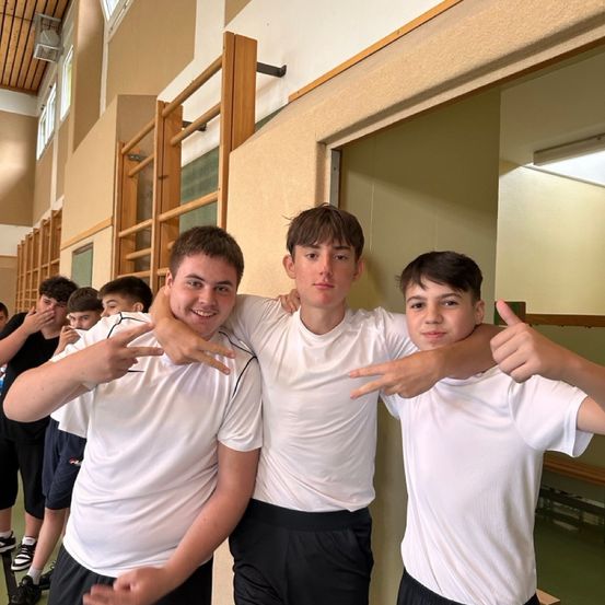 Drei Jungen in weißen T-Shirts und schwarzen Shorts posieren für ein Foto in einem Flur. Ein Junge gibt ein Daumen hoch, während die anderen Handgesten machen. Andere Kinder sind im Hintergrund.