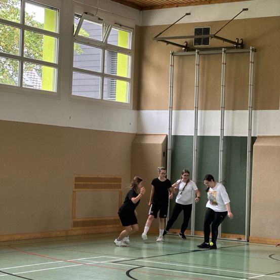 Vier Frauen spielen ein Spiel auf einem Indoor-Feld. Sie tragen Turnschuhe und Freizeitkleidung. Das Feld hat einen grünen Boden mit weißen Markierungen.