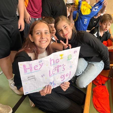 Zwei Mädchen halten ein Schild mit der Aufschrift Hey ho lass uns gehen, eines von ihnen macht ein Friedenszeichen. Sie sitzen mit anderen Kindern in einem Klassenzimmer.