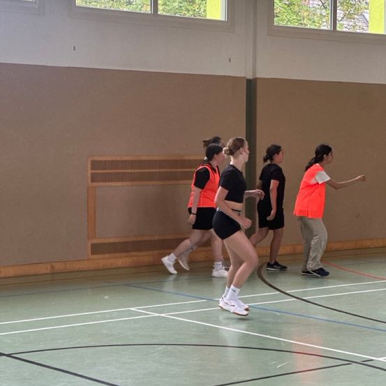 Fünf Frauen in Sportkleidung joggen an den Seitenlinien eines Hallenspielfelds, eine Frau zeigt. Die Wände sind braun und Fenster sind oben sichtbar.