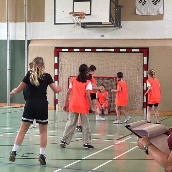 Eine Gruppe junger Frauen in orangefarbenen Westen spielt Basketball auf einem Innenfeld, eine hält ein Buch. Ein Tor ist im Hintergrund.