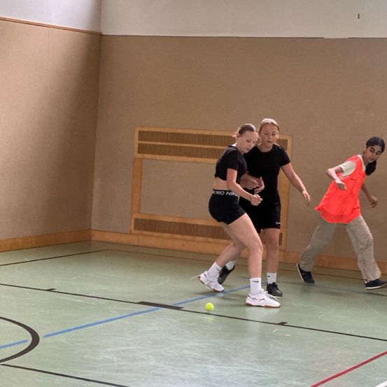 Drei Personen spielen Tennis in einer Halle. Eine Frau versucht, einen gelben Ball zu schlagen, während eine andere Frau in der Nähe steht. Der dritte Spieler in einem orangefarbenen Weste läuft auf sie zu.