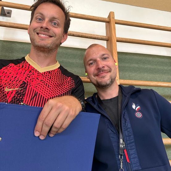 Zwei Männer stehen lächelnd, einer in einem rot-schwarzen Sporttrikot, der andere in einem marineblauen Jackett. Sie befinden sich in einer Indoor-Sportanlage mit Holzstangen im Hintergrund.