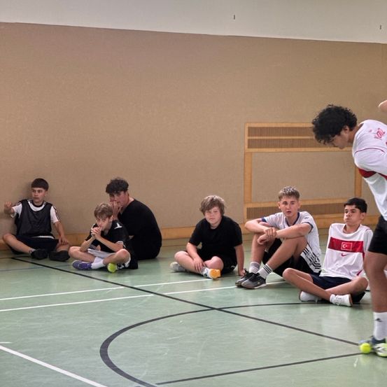 Mehrere junge Jungen in Sportkleidung sitzen auf dem Boden einer Indoor-Sporthalle, einige beobachten einen Mann mit einem Tennisschläger.