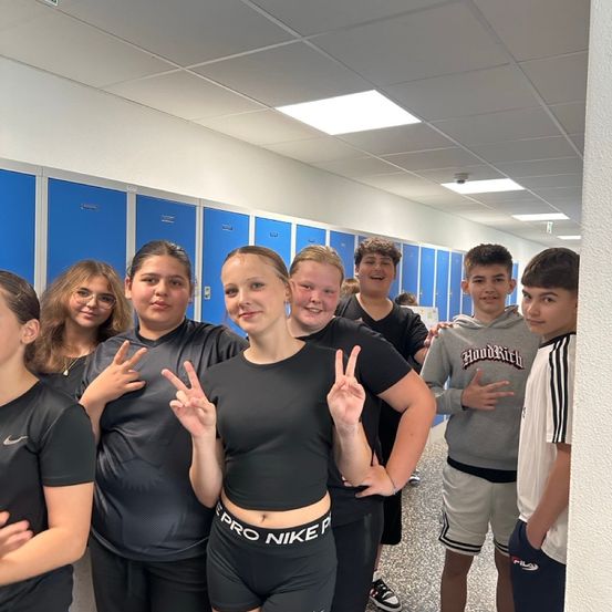 Eine Gruppe junger Leute steht in einem Flur mit blauen Schließfächern, lächelt und posiert für ein Foto. Ein Mädchen in der Mitte trägt ein schwarzes Pro Nike-Shirt und Shorts, während andere verschiedene farbige Shirts tragen.