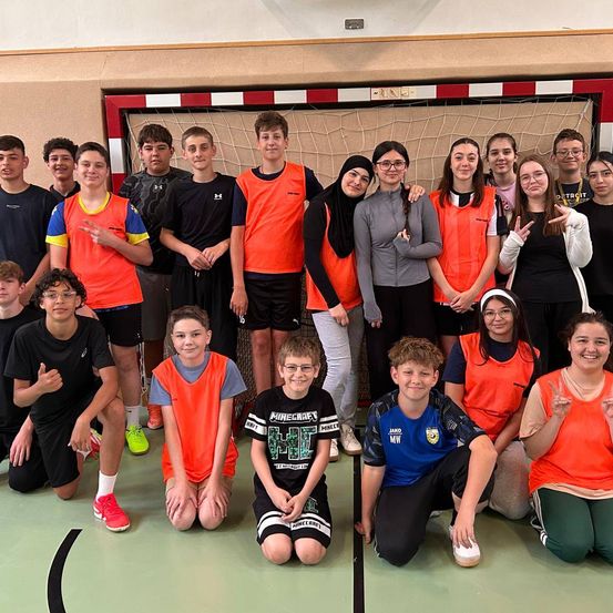 Eine Gruppe von Teenagern posiert für ein Foto in einer Turnhalle mit einem Fußballtor im Hintergrund, trägt orange Westen und lächelt.