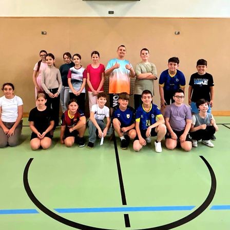 Eine Gruppe von Kindern und Erwachsenen posiert für ein Foto in einer Turnhalle, einige kniend und andere stehend. Sie tragen verschiedenfarbige T-Shirts und einige tragen eine Brille. Ein Basketballfeld ist im Hintergrund zu sehen.