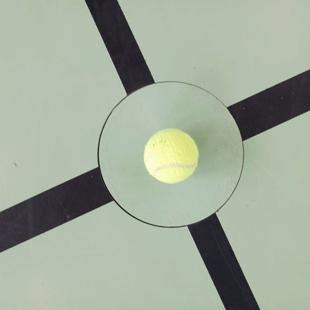 Ein Tennisball liegt in einem Glaskreis auf einer grünen Oberfläche mit schwarzen Linien, die ein Quadrat bilden.