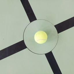 Ein Tennisball liegt in einem Glaskreis auf einer grünen Oberfläche mit schwarzen Linien, die ein Quadrat bilden.